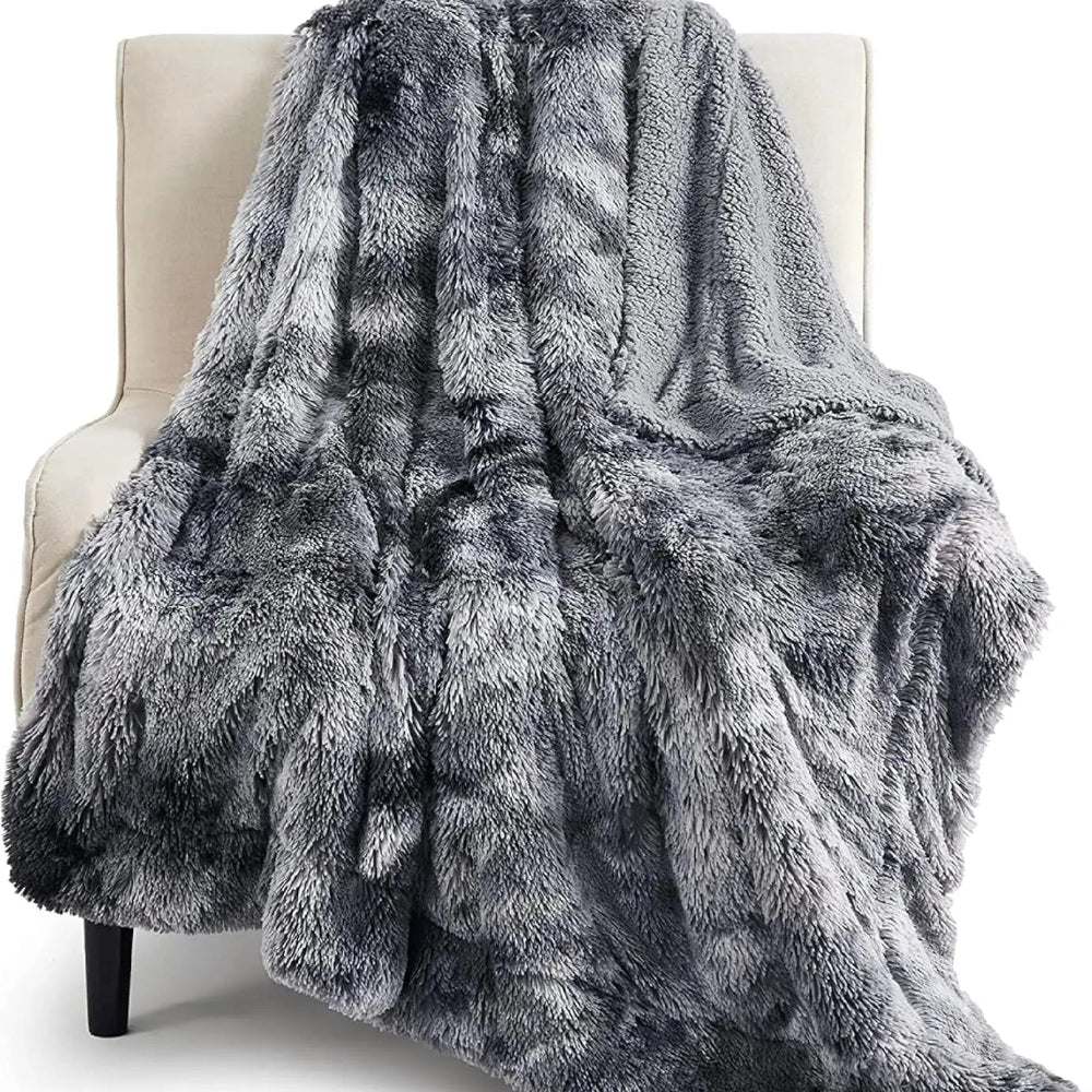 Un plaid en fausse fourrure gris ultra doux et épais, drapé sur un fauteuil clair, avec une texture moelleuse et un effet visuel chaleureux et cocooning.