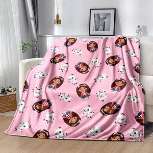 Un plaid enfant à imprimé Gabby Dollhouse recouvrant un canapé, avec des personnages ludiques répétés sur toute la surface, créant une ambiance douce et joyeuse dans un intérieur lumineux.