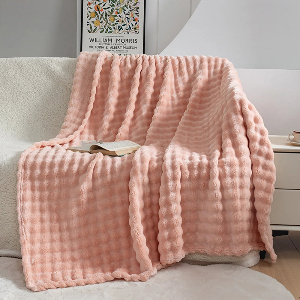 Un plaid fausse fourrure doux et moelleux à texture gaufrée, délicatement drapé sur un fauteuil avec un livre posé dessus, créant une atmosphère chaleureuse et invitante dans un salon lumineux.