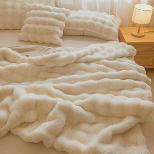 Un plaid fausse fourrure ultra doux au relief ondulé, délicatement posé sur un lit avec des coussins assortis, créant une ambiance chaleureuse et cocooning dans une chambre lumineuse.