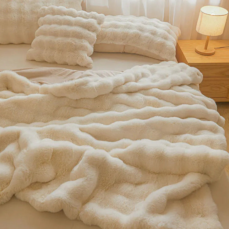 Un plaid fausse fourrure ultra doux au relief ondulé, délicatement posé sur un lit avec des coussins assortis, créant une ambiance chaleureuse et cocooning dans une chambre lumineuse.