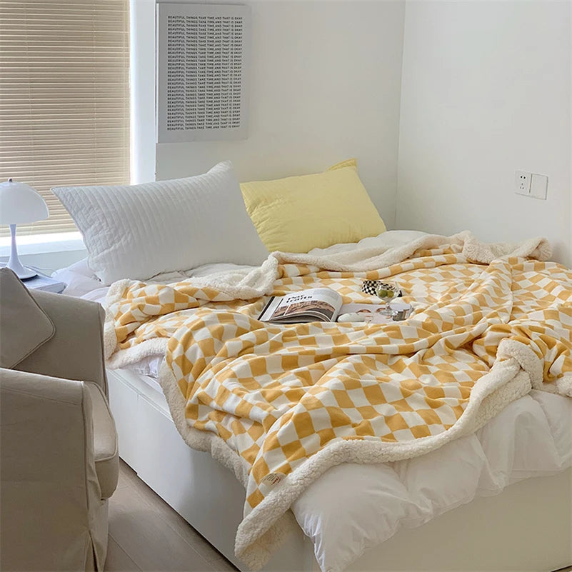 Un plaid polaire motif damier avec bordure duveteuse posé sur un lit lumineux, légèrement froissé pour créer une ambiance cocooning.