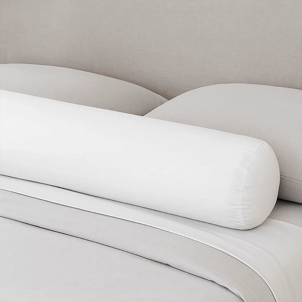 Un traversin de lit en velours moelleux de forme cylindrique, posé sur un lit aux draps clairs devant une tête de lit beige, créant une atmosphère douce et minimaliste dans la chambre.