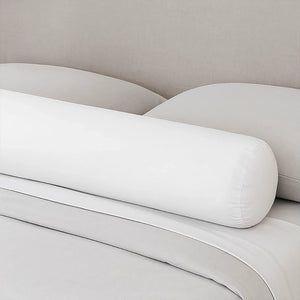 Un traversin de lit en velours moelleux de forme cylindrique, posé sur un lit aux draps clairs devant une tête de lit beige, créant une atmosphère douce et minimaliste dans la chambre.