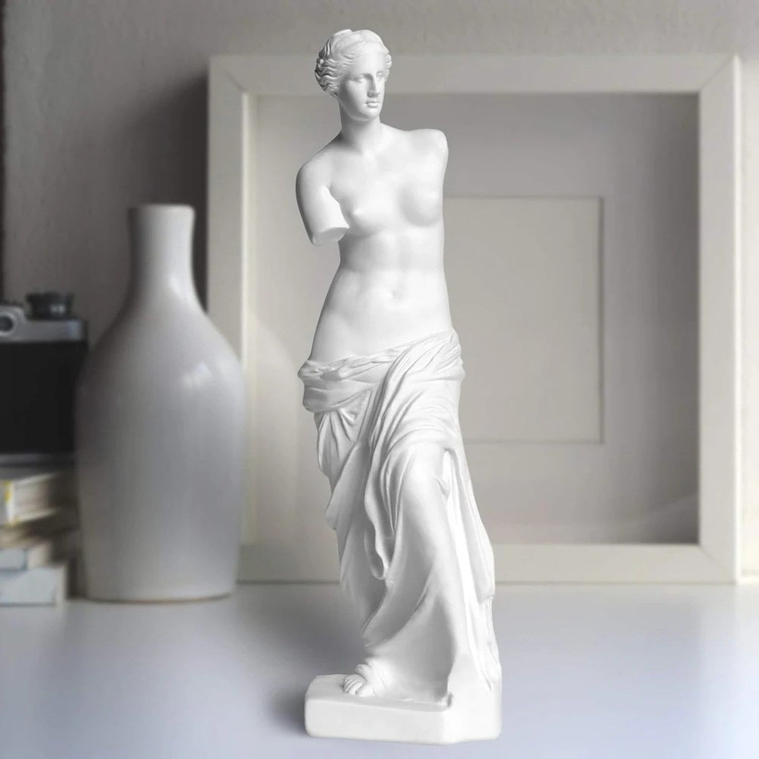 Sculpture Vénus de Milo posée sur une surface blanche avec vase et cadre en arrière-plan flou