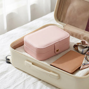 Boite à bijoux rose fermée dans une valise ouverte sur un lit avec draps blancs et foulard