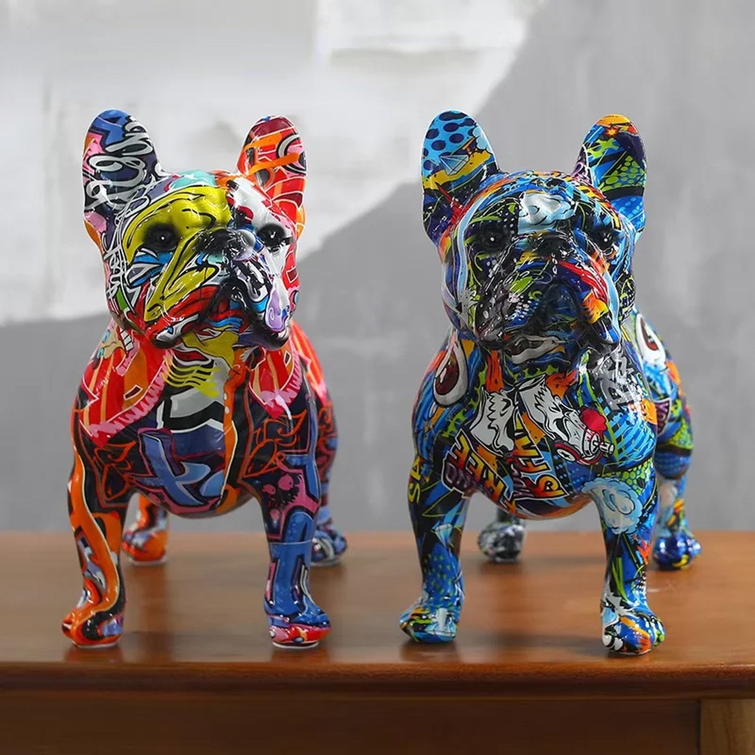Deux statues bouledogue français en résine avec motifs graffiti multicolores posées sur une table en bois