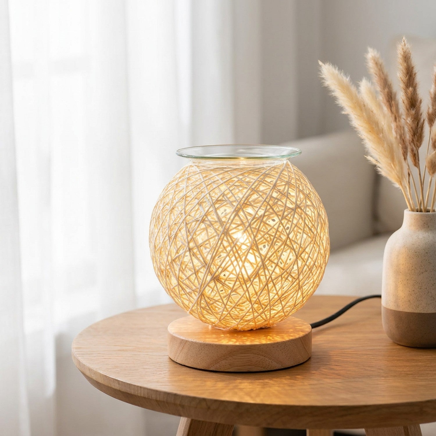 Brûle-parfum en rotin tressé sur une table ronde près d'une fenêtre avec des pampas séchées en lumière douce