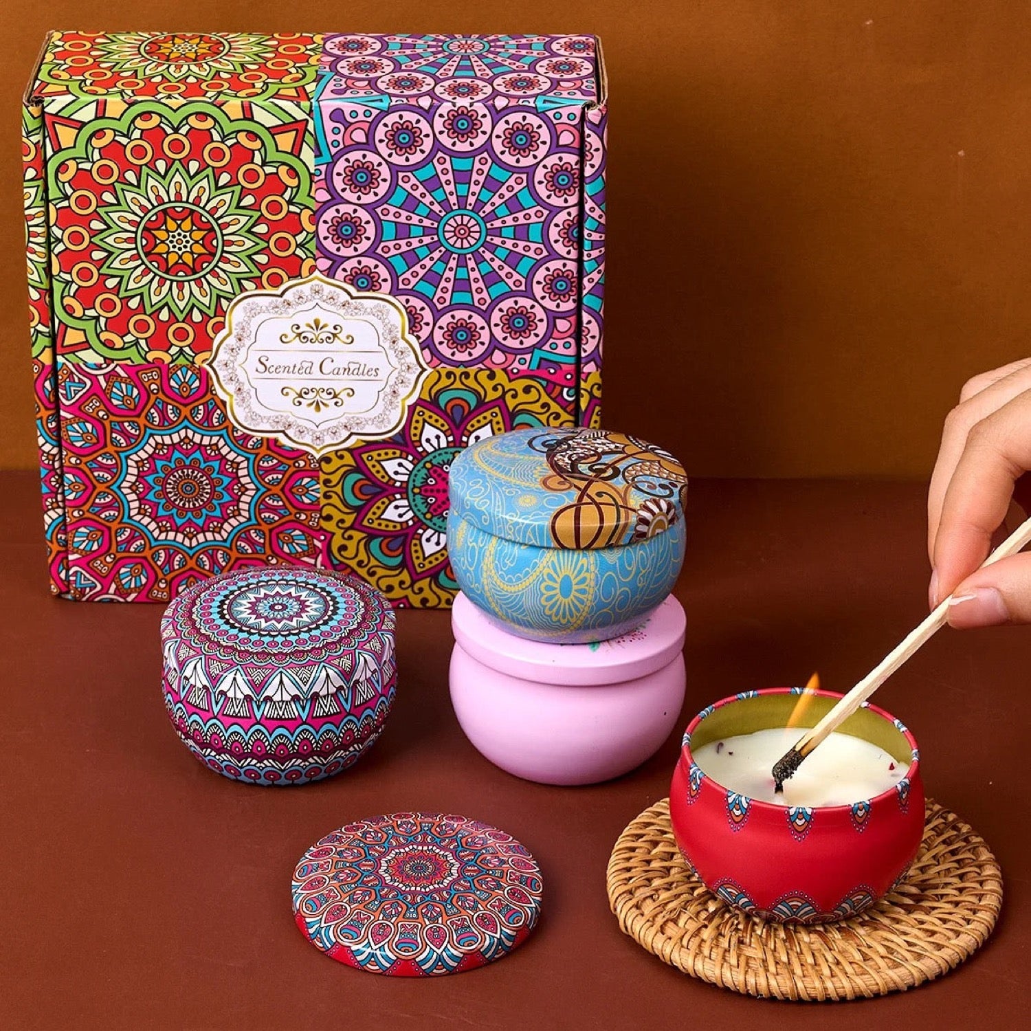 Coffret cadeau mandala coloré avec quatre bougies parfumées en boîtes métalliques décorées et une main allumant une bougie sur un dessous de verre en osier
