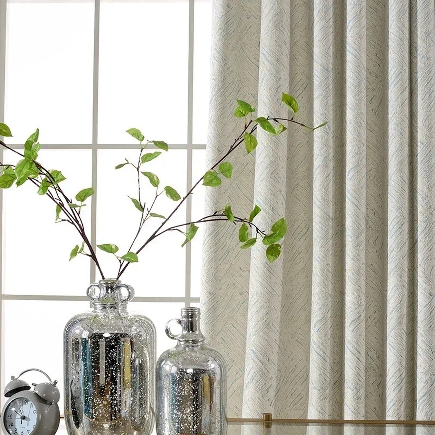 Détail du rideau chenille devant une fenêtre avec vase argenté et branches vertes