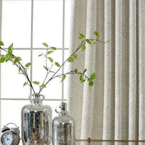 Détail du rideau chenille devant une fenêtre avec vase argenté et branches vertes