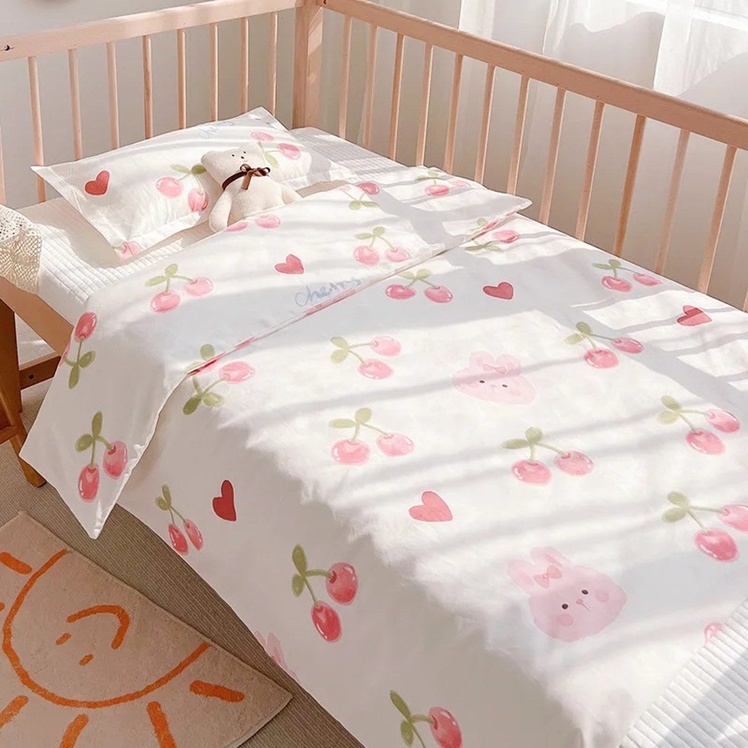 Parure de lit bébé motif lapin cerise installée sur un lit à barreaux avec un nounours