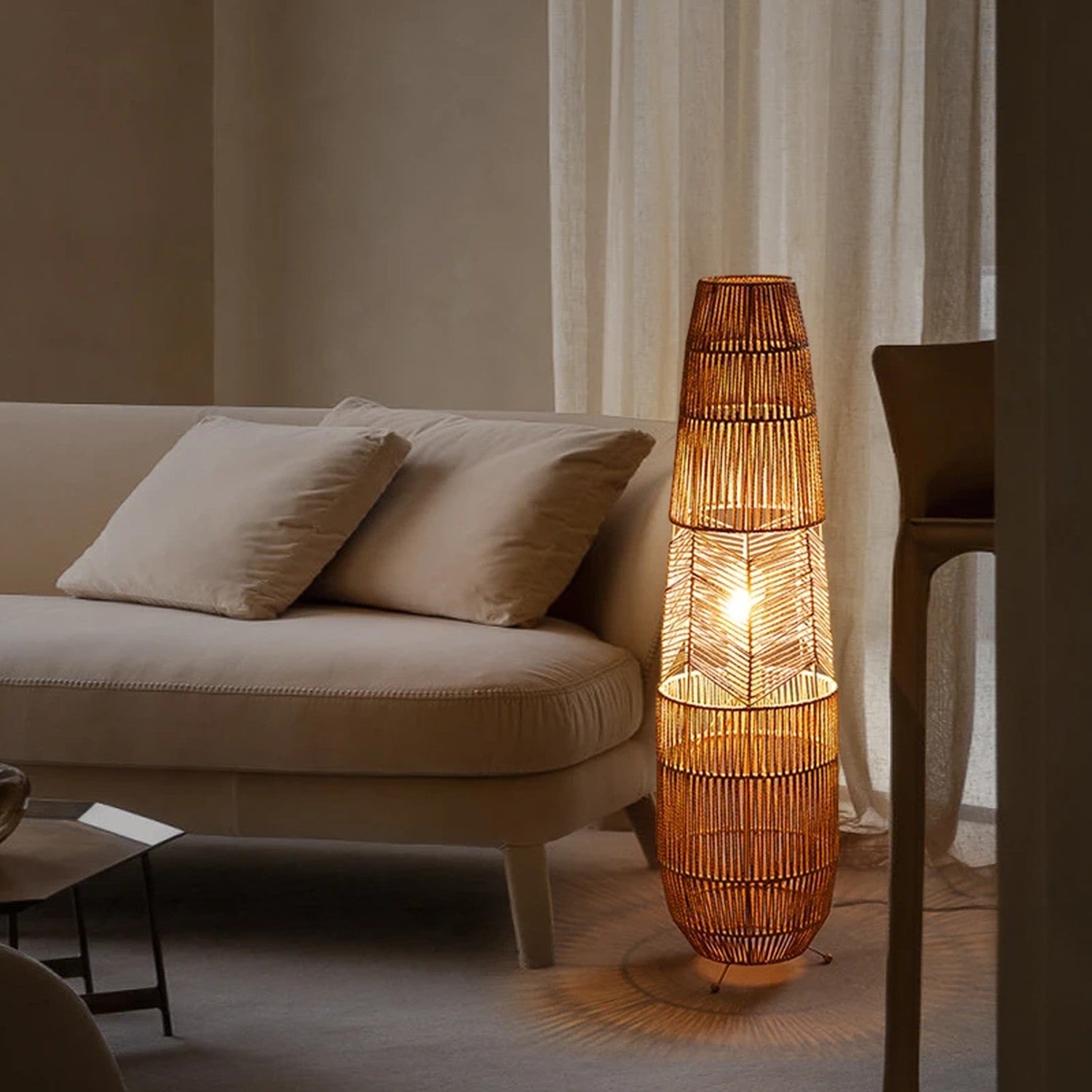 Lampadaire en rotin tressé diffusant une lumière chaude devant un canapé et des rideaux