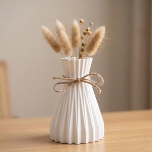 Vase origami plissé blanc en plastique avec un nœud en jute et des fleurs séchées posé sur une étagère