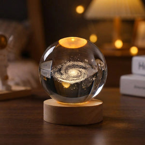 Boule de cristal gravée en 3D représentant une galaxie éclairée d'une lumière chaude sur socle en bois