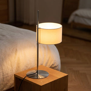 Lampe à poser design japonais en métal chrome avec abat-jour cylindrique blanc allumée sur une table de chevet