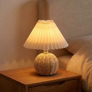 Lampe de chevet à base en rotin tressé et abat-jour plissé blanc allumée sur une table de nuit