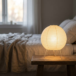 Lampe wabi-sabi en papier de riz allumée sur une table de chevet en bois près d'un lit en lin