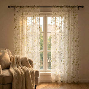 Voilage fleuri vert baigné de lumière dorée près d'un fauteuil cocooning et d'un plaid