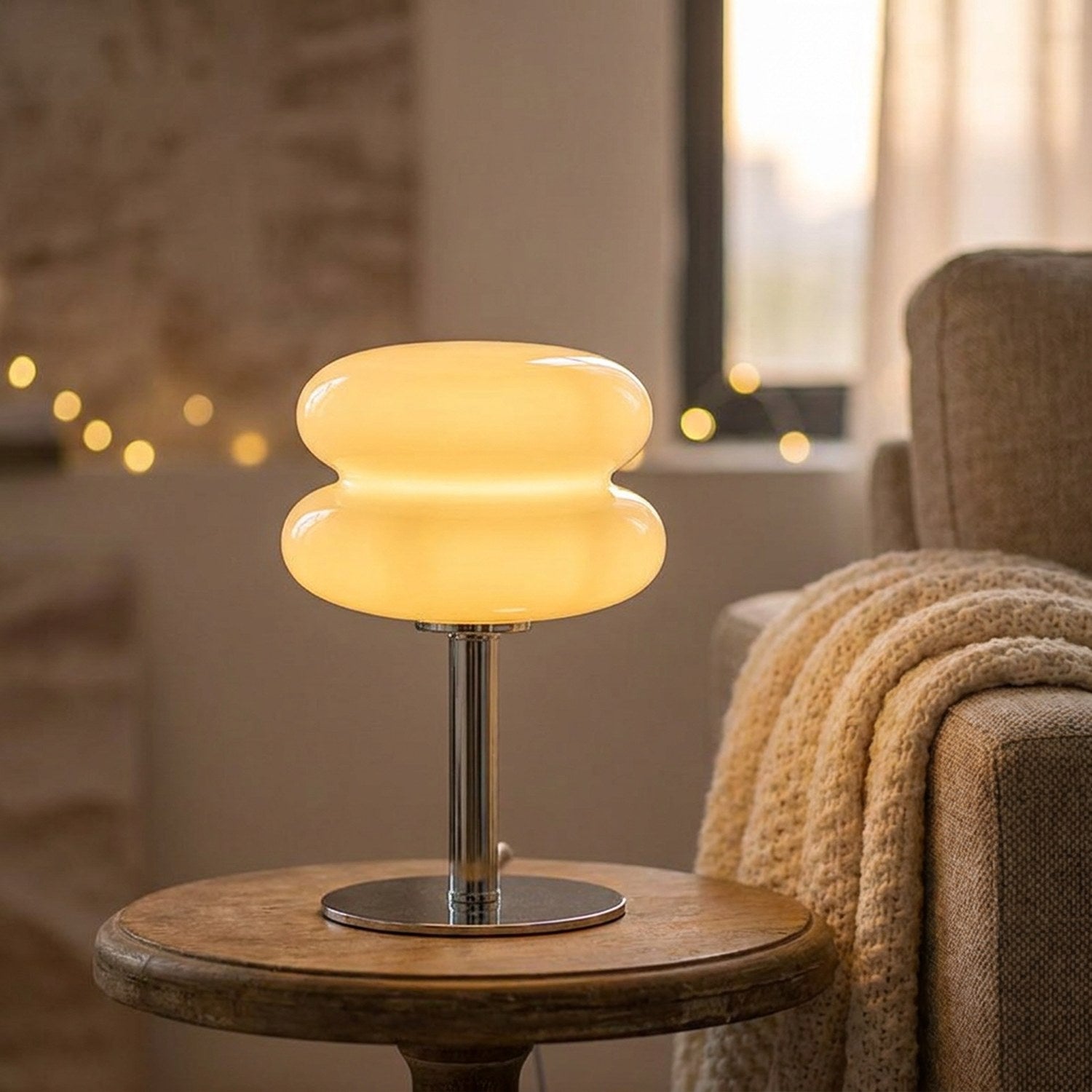 Lampe de chevet en verre forme macaron allumée sur une table basse en bois près d'un fauteuil avec un plaid