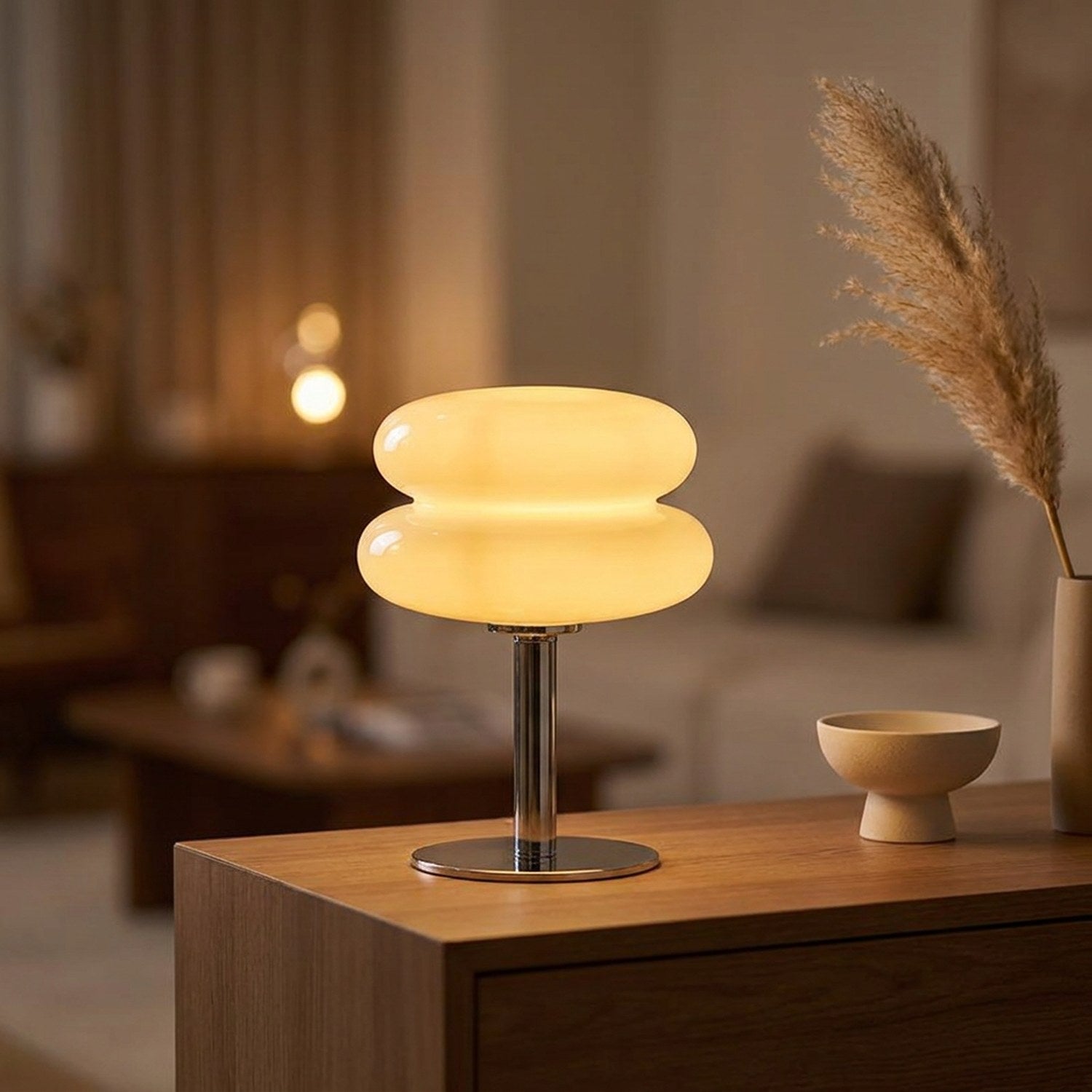 Lampe de chevet en verre forme macaron allumée sur une console en bois avec une plume de pampa