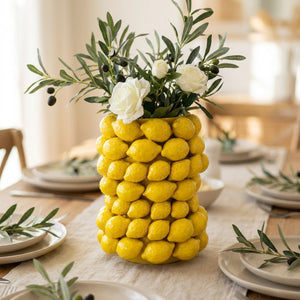 Vase citron en céramique jaune au centre d'une table d'été dressée avec un chemin de table en lin et des branches d'olivier