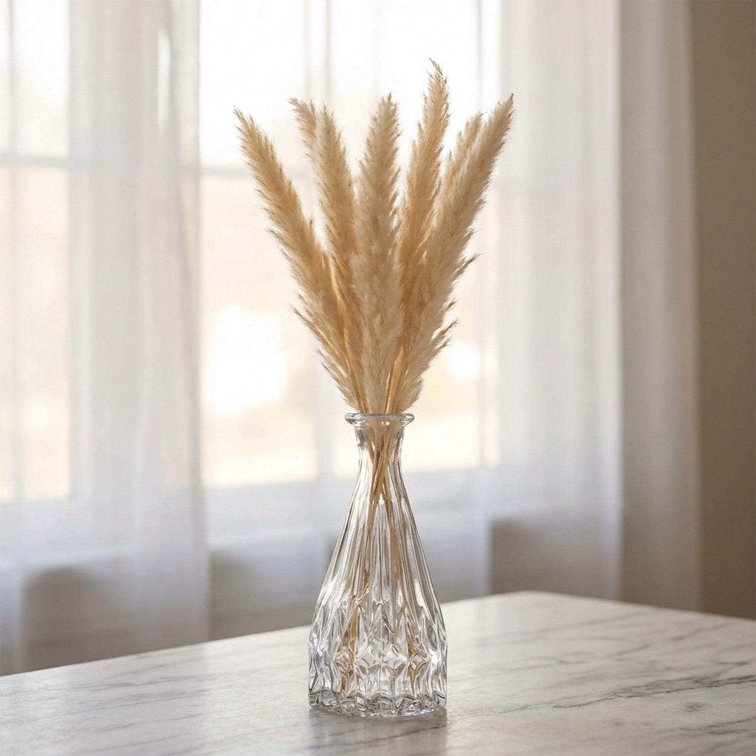 Vase en verre vintage à facettes avec des pampas séchées posé sur un plan en marbre devant une fenêtre voilée