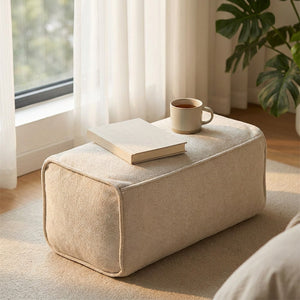 Pouf repose-pieds beige avec un livre et une tasse près d'une fenêtre lumineuse