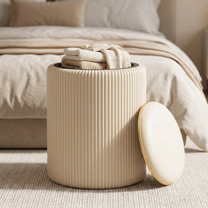 Tabouret de rangement beige ouvert au pied d'un lit révélant des plaids rangés