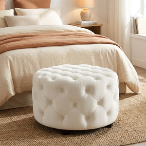 Tabouret pouf blanc en velours posé au pied d'un lit dans une chambre chaleureuse