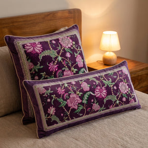 Coussins carré et rectangulaire en velours violet à motif floral disposés sur un lit avec lampe de chevet chaleureuse