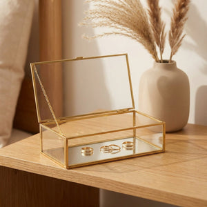 Boîte à bijoux en verre ouverte sur une table en bois avec pampa et bagues