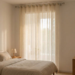 Voilage tulle beige suspendu devant une fenêtre de chambre avec lit en lin naturel