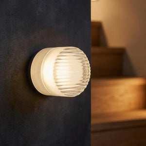 Applique murale LED blanche côtelée sur un mur sombre près d'un escalier en bois