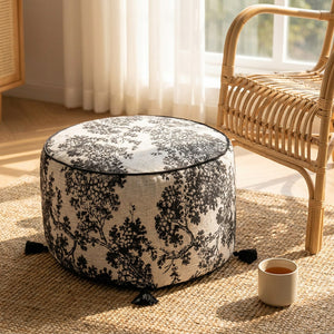 Pouf rond toile de Jouy posé sur un tapis en jute près d'un fauteuil en rotin