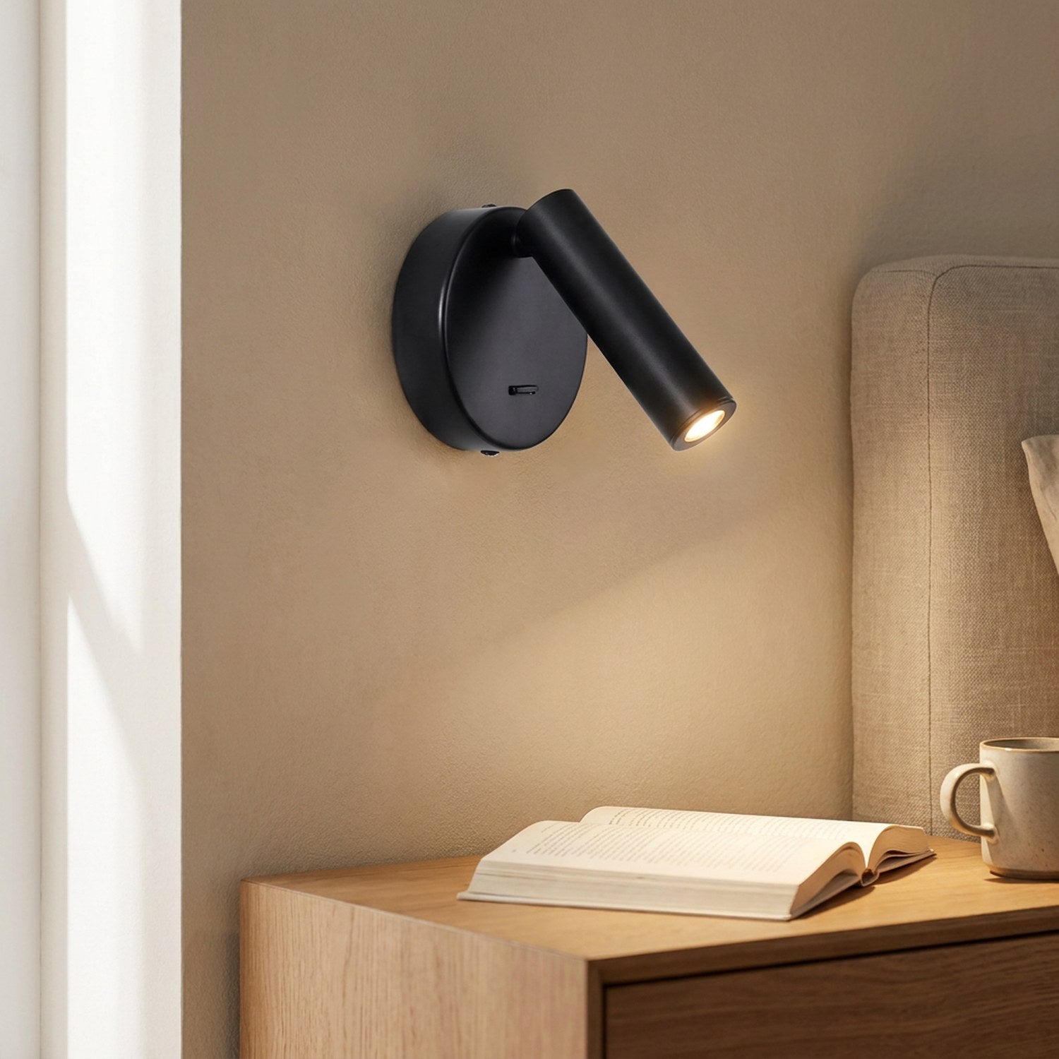Applique murale ronde noire au spot LED orientable allumée au-dessus d'une table de chevet en bois avec un livre ouvert
