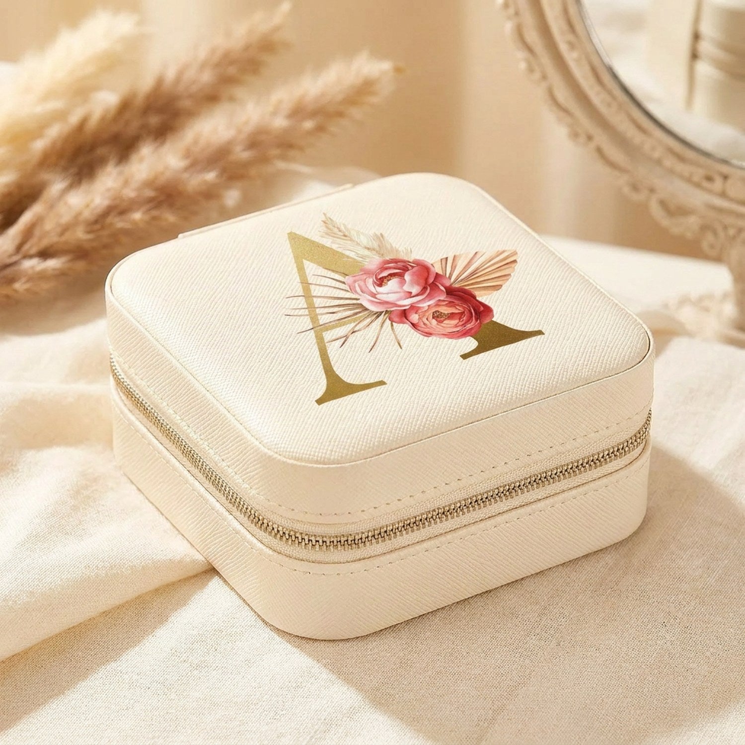 Boîte à bijoux de voyage crème à initiale dorée et fleurs bohème posée sur une coiffeuse