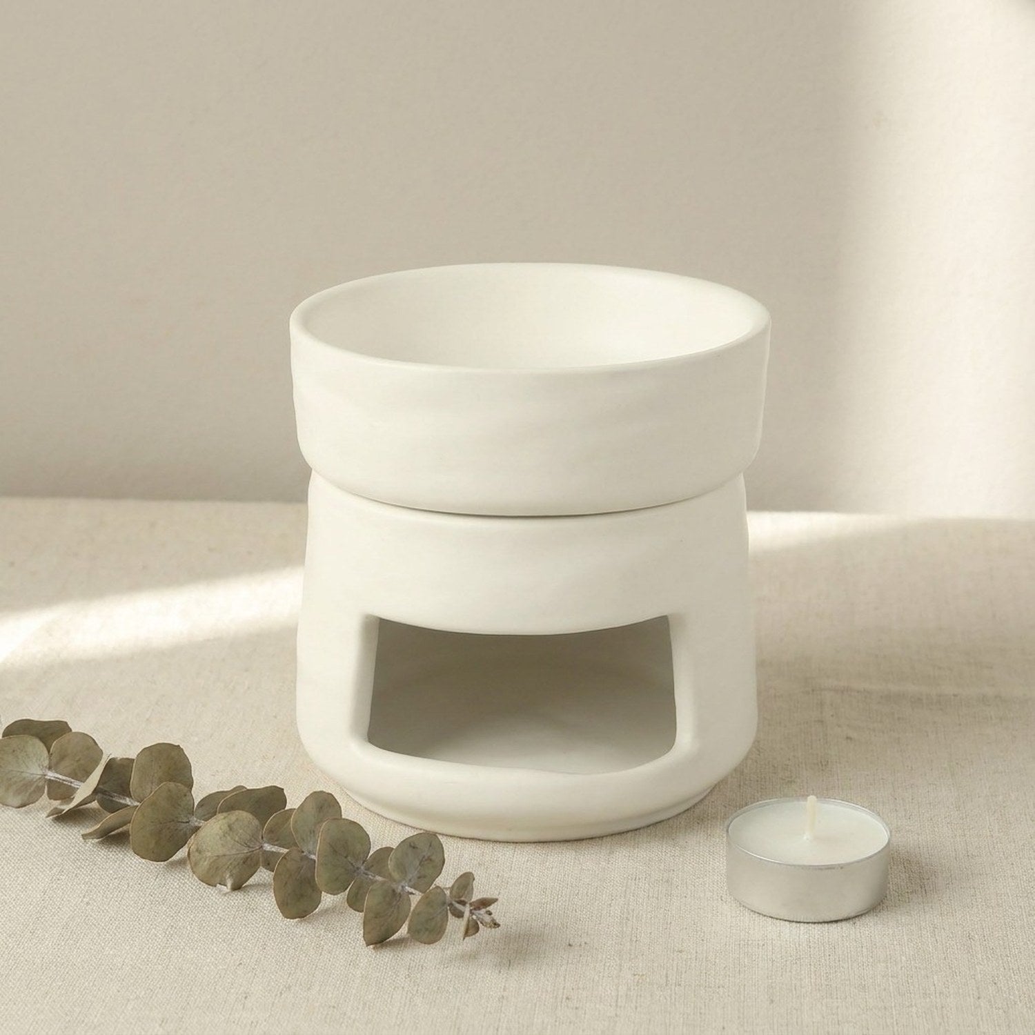 Brûle-parfums en céramique mate blanche posé sur surface en lin avec brindilles d'eucalyptus