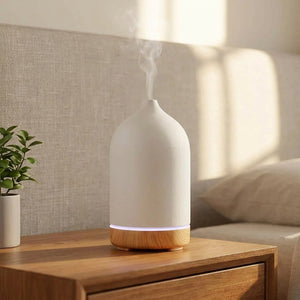 Diffuseur d'huiles essentielles en céramique blanche à base bois diffusant une brume sur une table de chevet