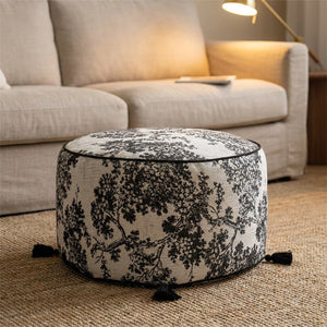Pouf rond en tissu jacquard motif toile de Jouy noir et blanc posé au sol devant un canapé beige
