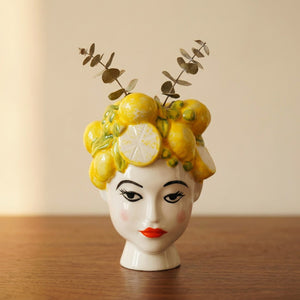 Vase en porcelaine représentant un visage de femme aux lèvres rouges couronné de citrons jaunes sur un meuble en bois