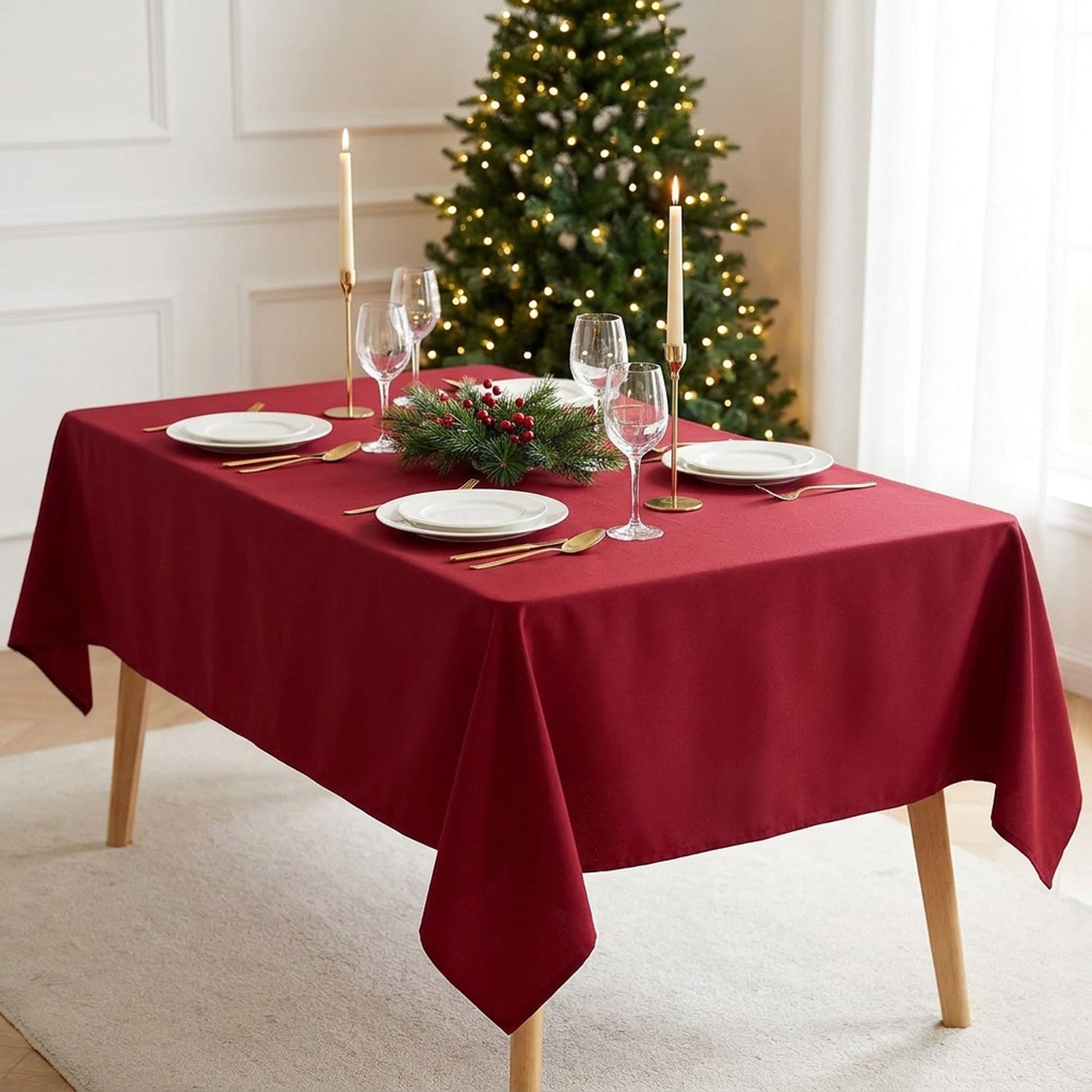 Nappe de Noël bordeaux unie sur une table rectangulaire dressée avec assiettes blanches, bougies dorées et sapin illuminé en arrière-plan