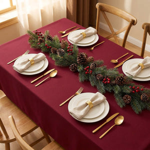 Vue plongeante d'une table de fêtes couverte de la nappe bordeaux avec quatre couverts dorés, serviettes en lin et guirlande de sapin centrale