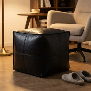 Pouf cube en cuir noir utilisé comme repose-pieds devant un fauteuil de lecture