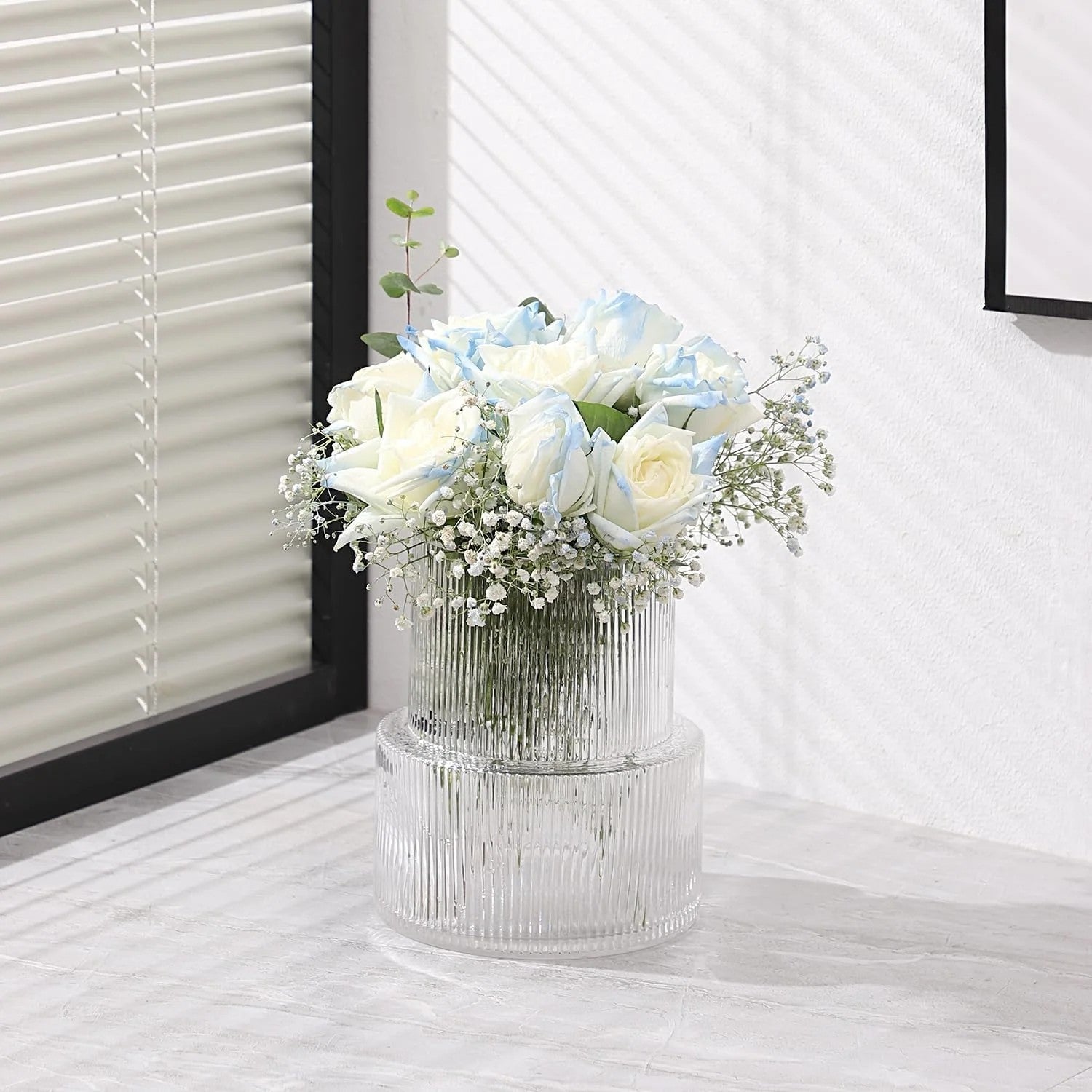 Vase en verre rayé avec roses bleues et blanches près d'une fenêtre à store blanc