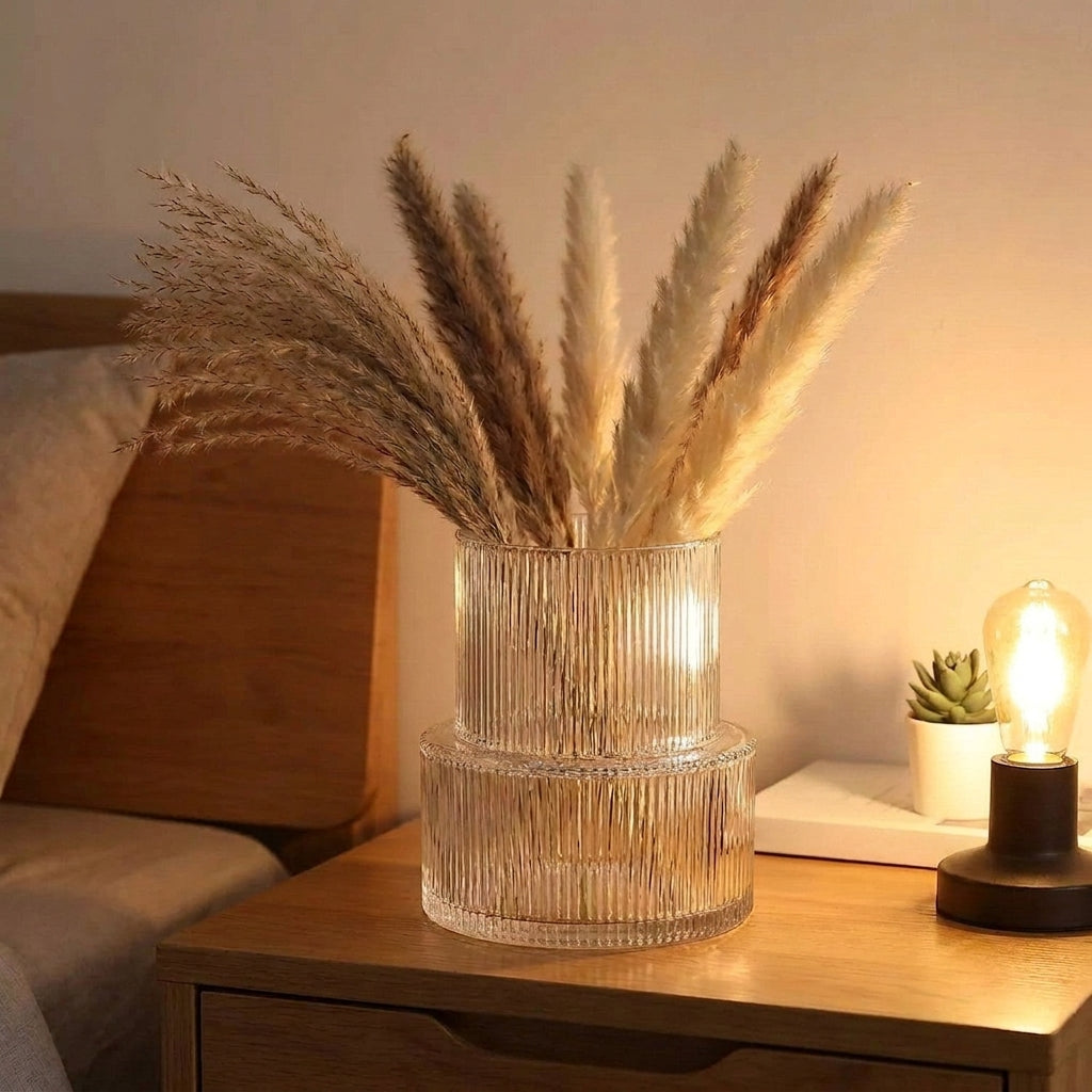 Vase en verre rayé avec herbes de pampa posé sur table de chevet dans une chambre au éclairage chaleureux