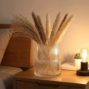 Vase en verre rayé avec herbes de pampa posé sur table de chevet dans une chambre au éclairage chaleureux