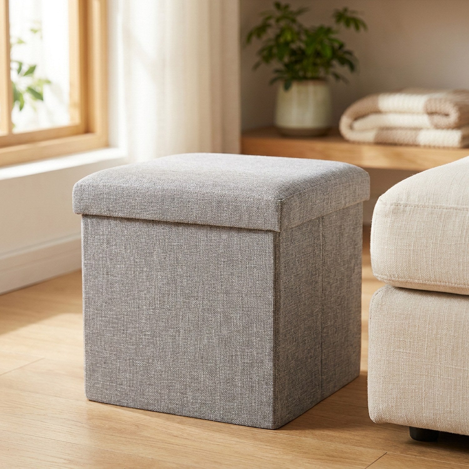 Tabouret de rangement pliable gris en tissu avec assise capitonnée posé dans un salon lumineux