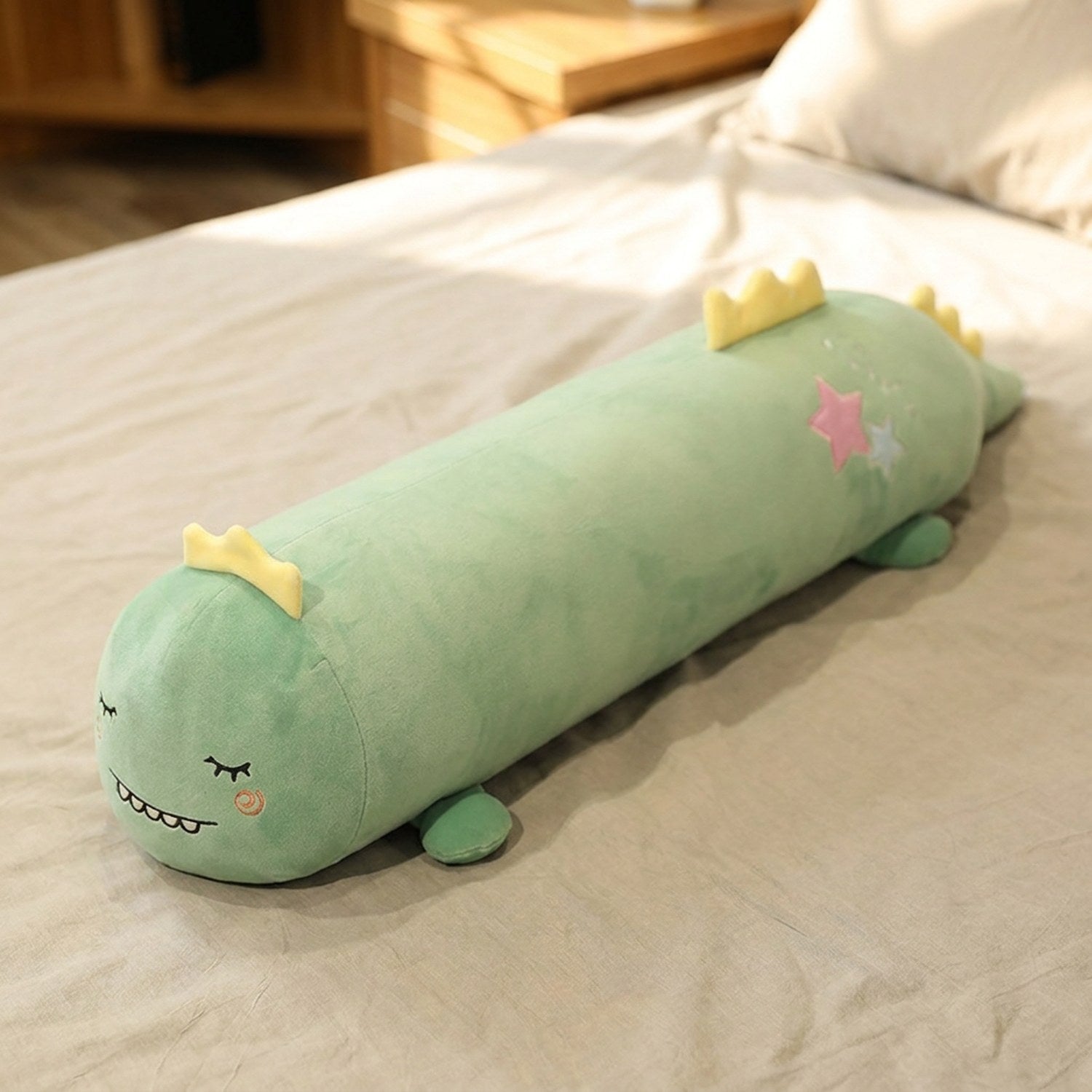Traversin peluche en forme de dinosaure kawaii posé sur un lit clair