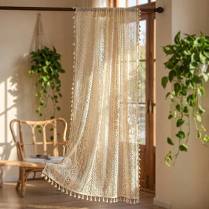 Rideau boho en dentelle au crochet beige à pompons suspendu devant une fenêtre lumineuse